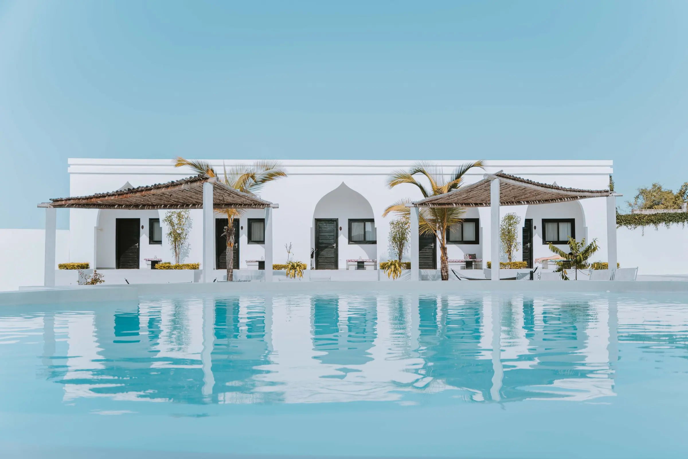 A low whitewashed villa with arched doorways and palm trees, reflected in a calm turquoise pool