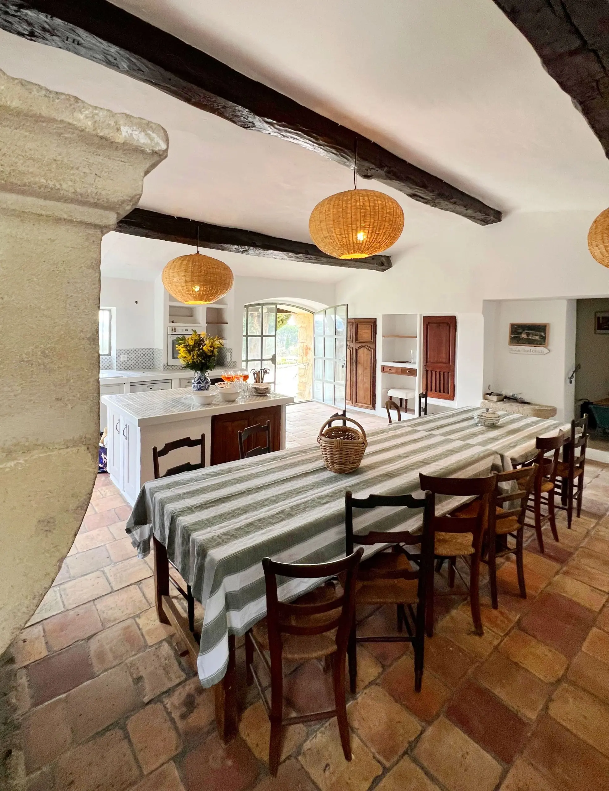 A whitewashed kitchen with timber beams, rattan lamps, terracotta tiles and a long table