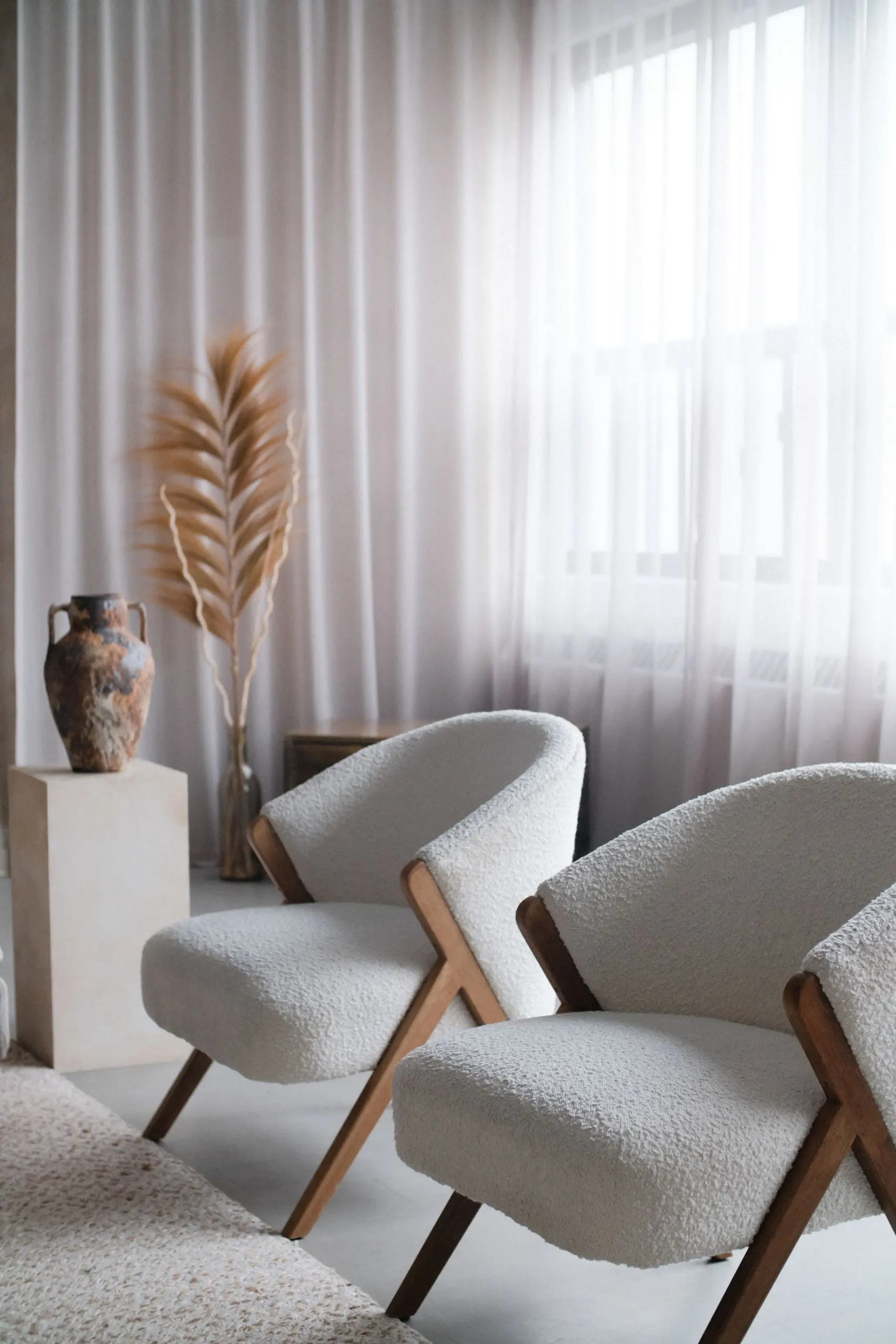 Two white armchairs before sheer linen curtains, with an old clay amphora and a dried palm leaf