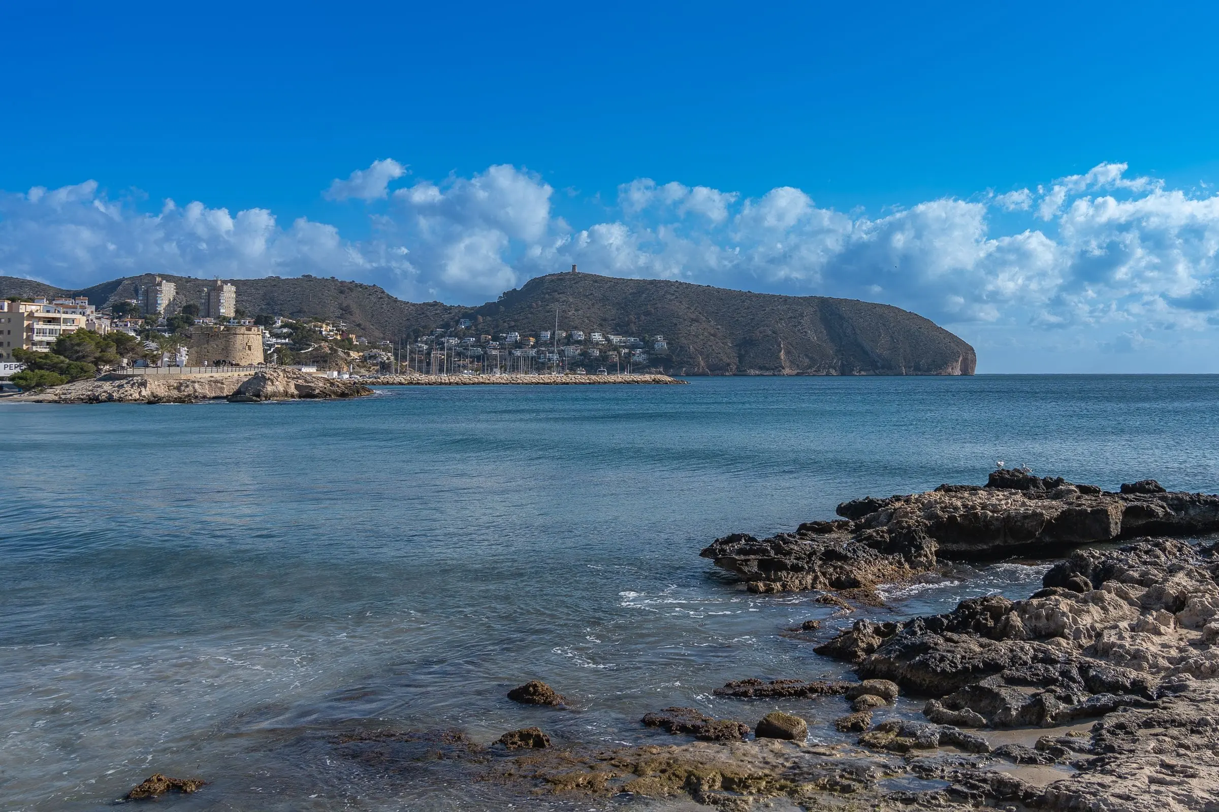 The rocky shore of Moraira with calm water and the Cap d’Or headland in the distance