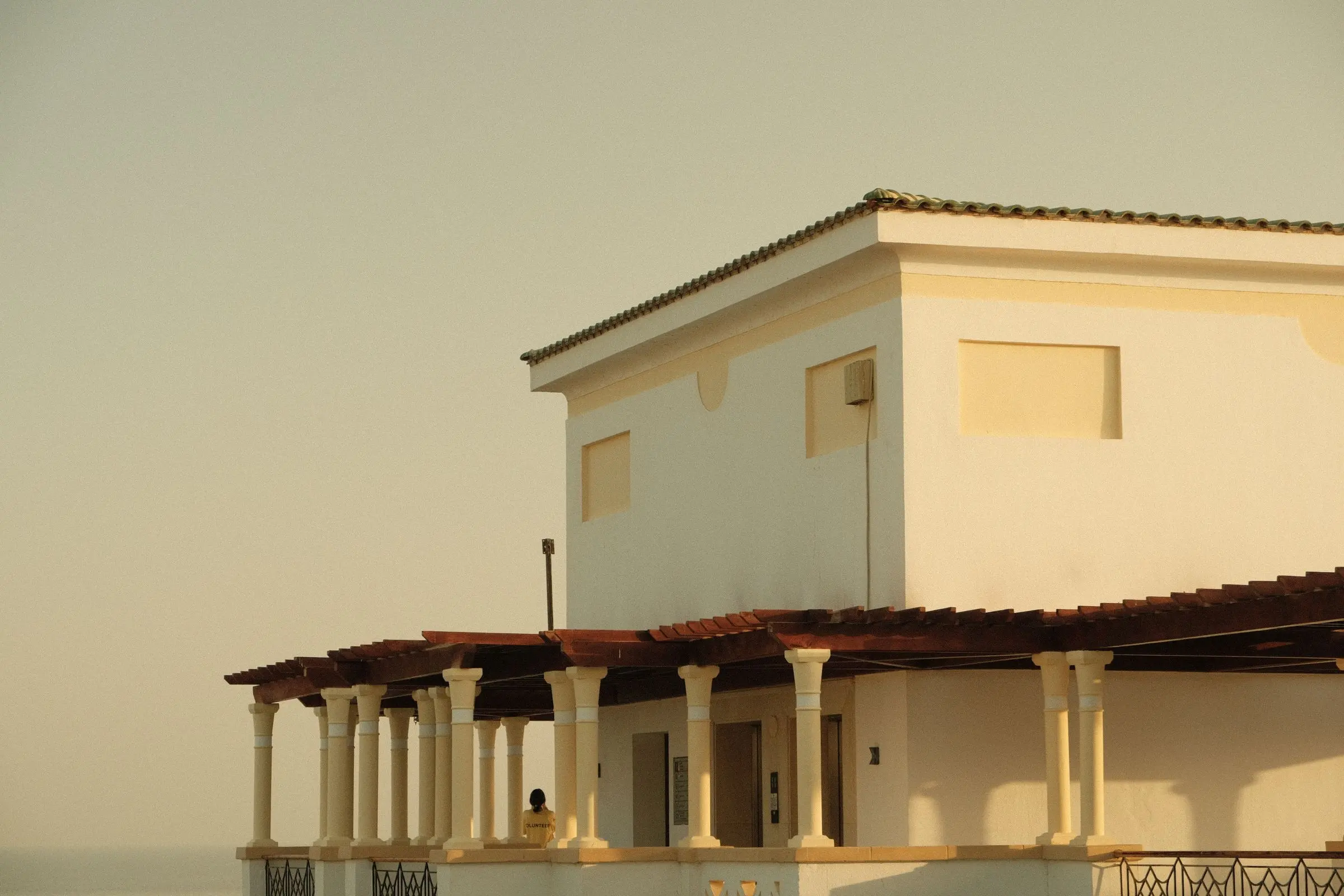 A whitewashed Mediterranean villa with a tiled roof and a colonnaded terrace in warm, hazy evening light